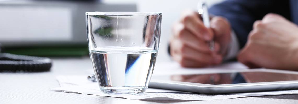 Glass of water on a table next to an ipad and someone writing in the background